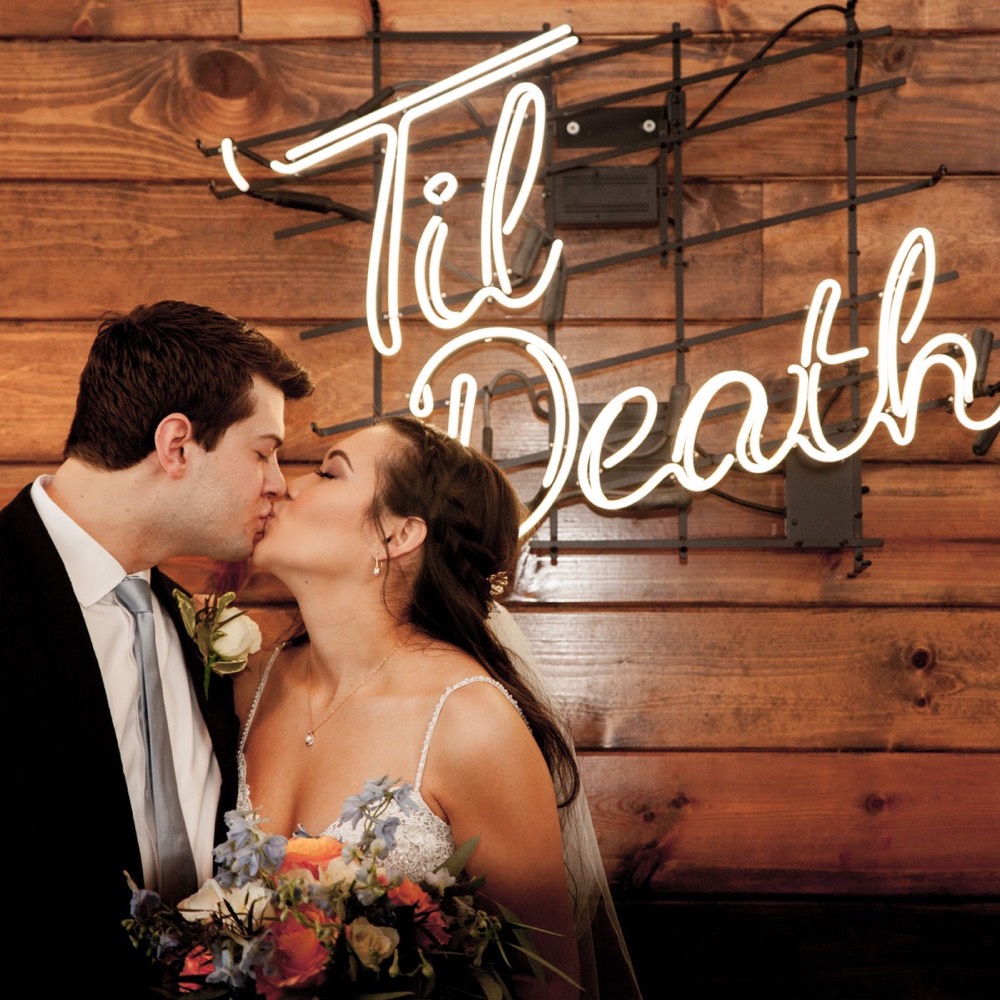 A couple kissing beneath a neon Til Death sign